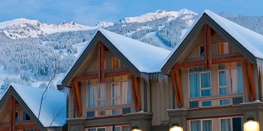 Snow-covered gables of timber lodge buildings, icicles at the eaves, snowy peaks behind.