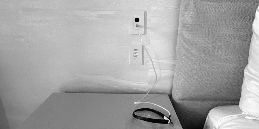 An oxygen outlet above a lighting keypad beside an upholstered headboard; a clear tube runs from the outlet to a black device on the nightstand.