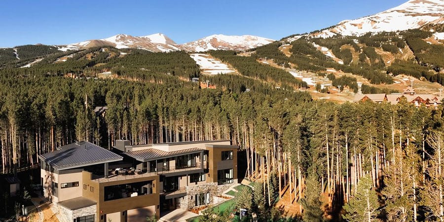 A contemporary mountain residence among dense evergreens, seen from above, with ski runs on the peaks beyond.