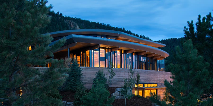 A glass and timber house among pines at twilight, its interiors lit warm against a blue sky.